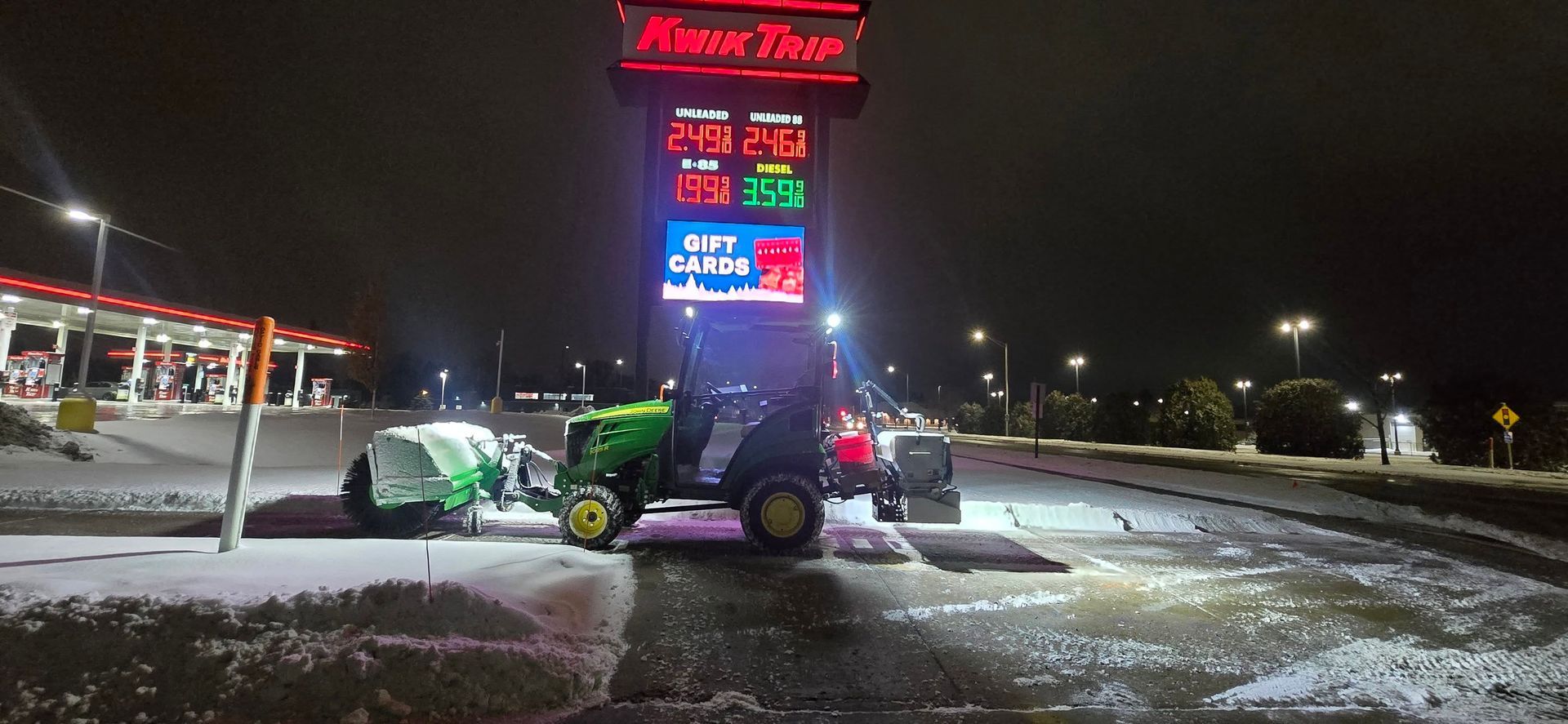 A tractor plowing snow at a gas station at night. Prices are displayed on a sign.