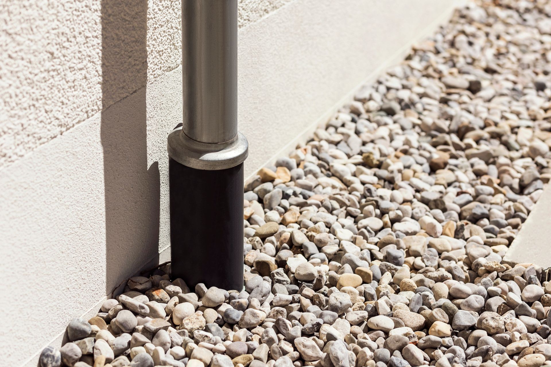 Silver downspout, black base, meets gravel bed beside a light-colored stucco wall in Wausau, WI.