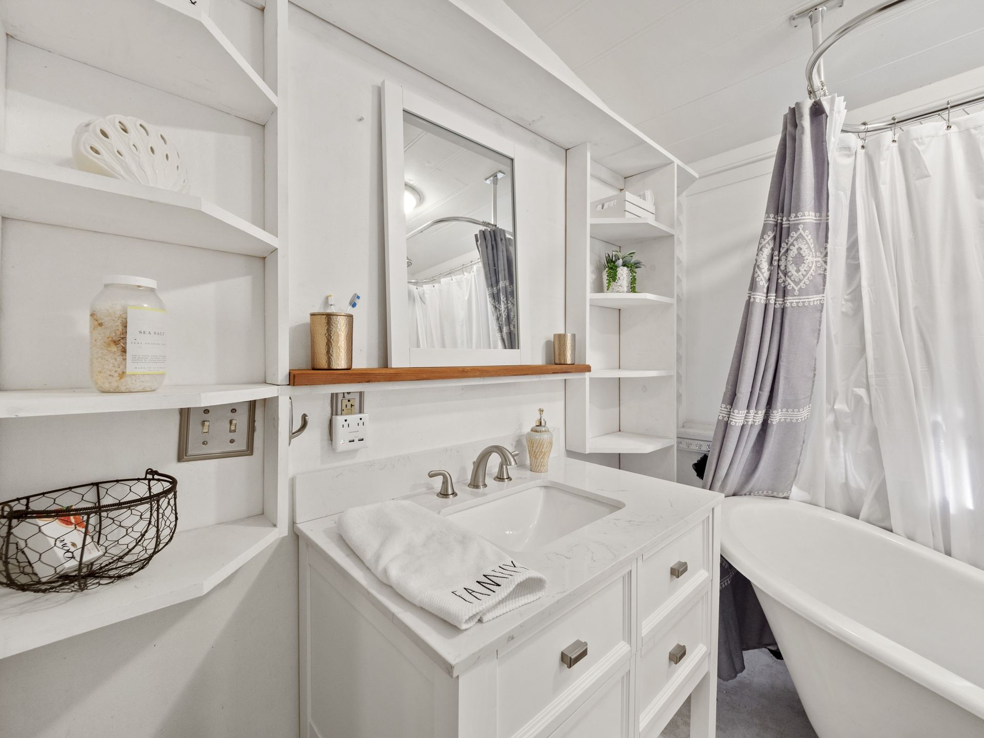 A bathroom with a sink , mirror , tub and shelves.
