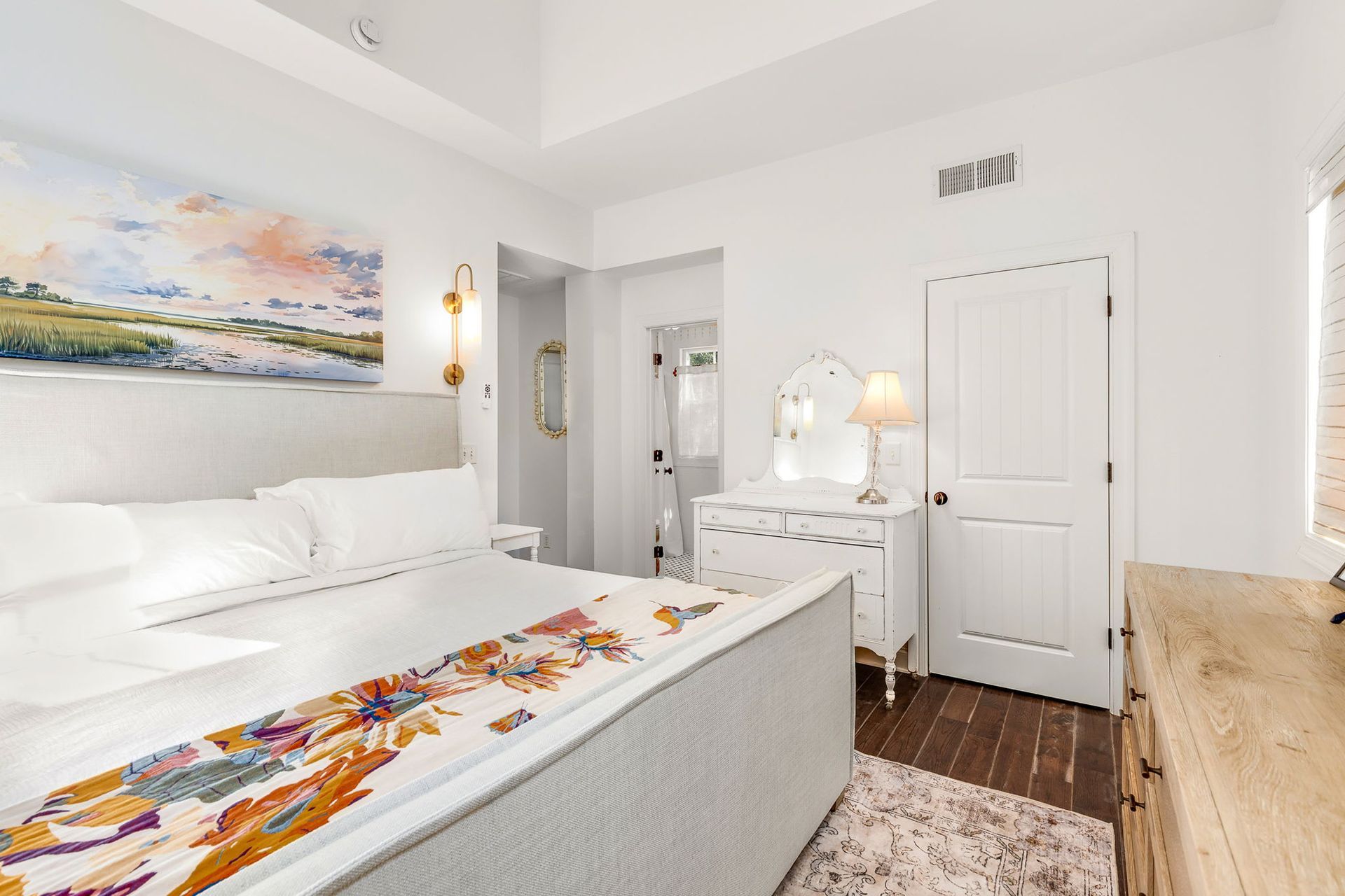 A bedroom with a large bed , dresser , mirror and painting on the wall.