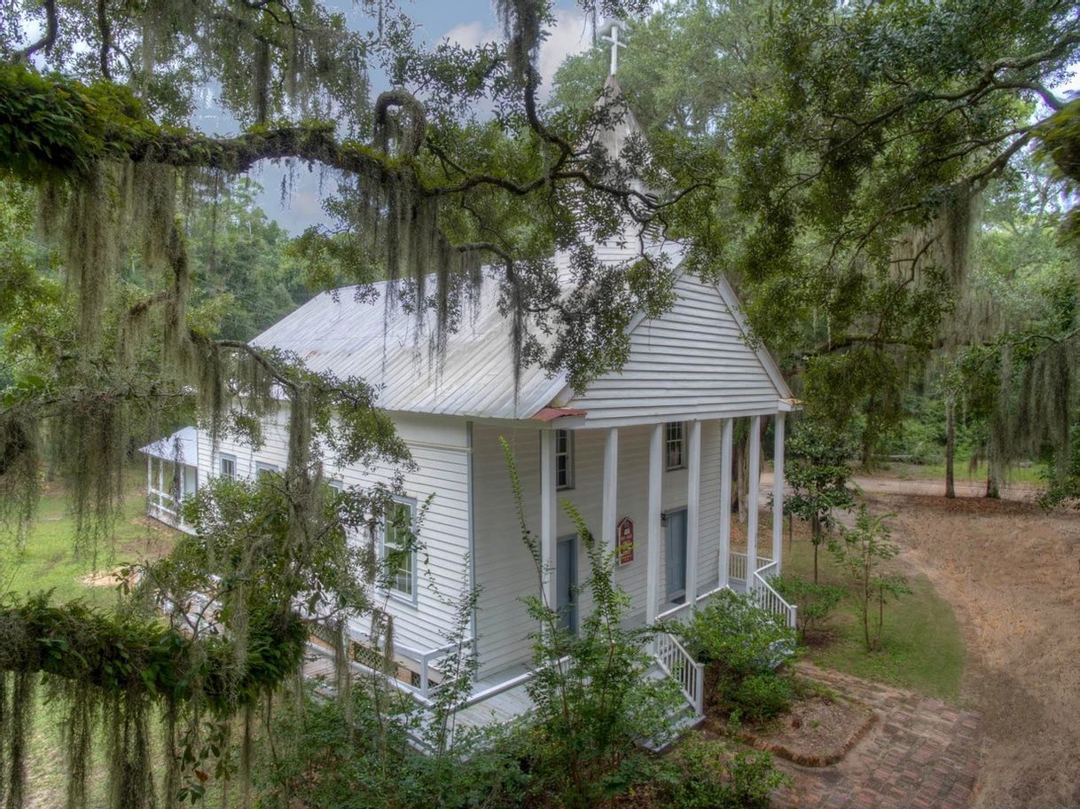 A white house with a porch is surrounded by trees and a dirt road.