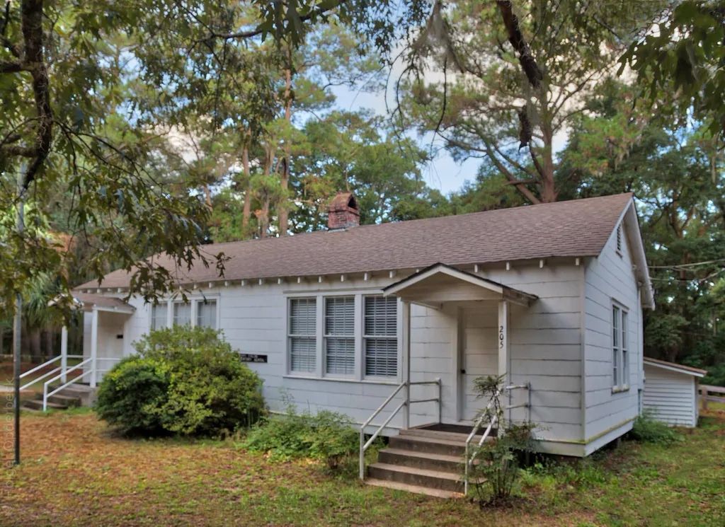 A small white house with a brown roof is surrounded by trees.