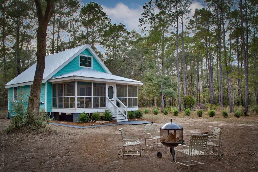 A small blue house with a screened in porch is in the middle of a forest.