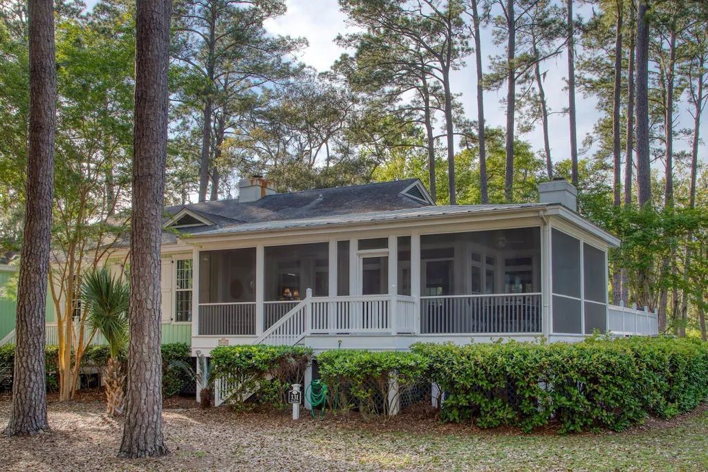 A small white house with a screened in porch is surrounded by trees.