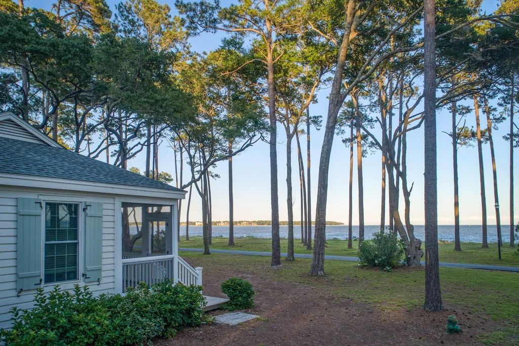 A small white house is surrounded by trees and a body of water.