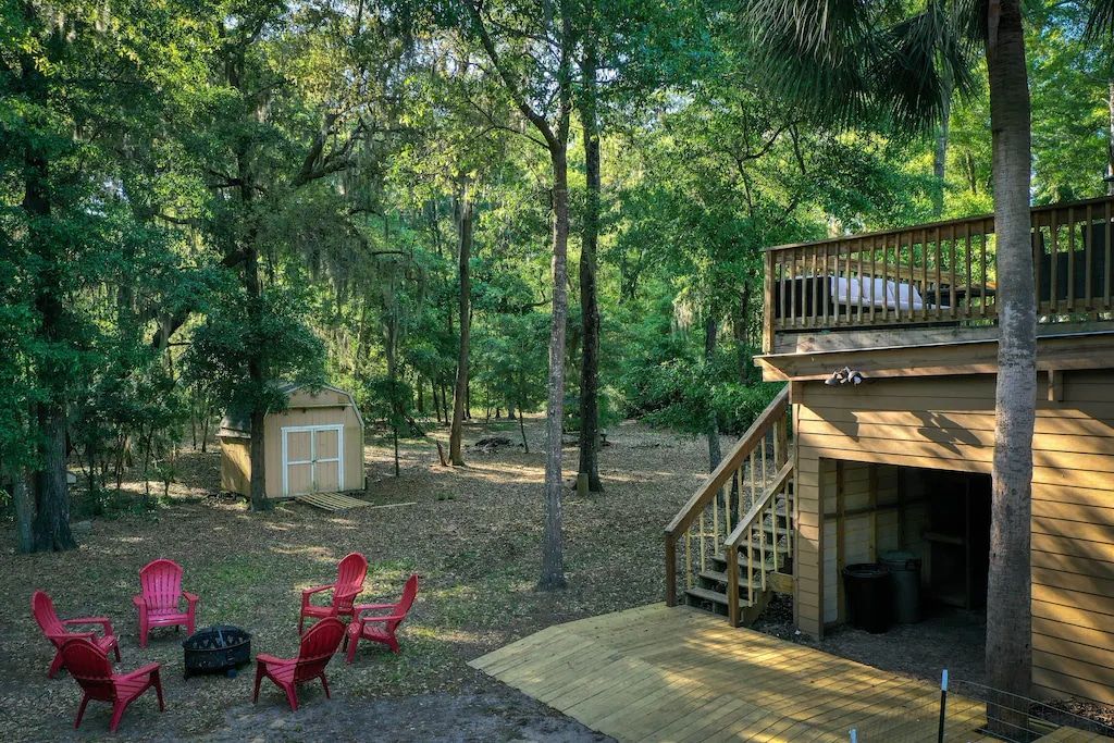A wooden deck with red chairs and a fire pit in front of a house in the woods.