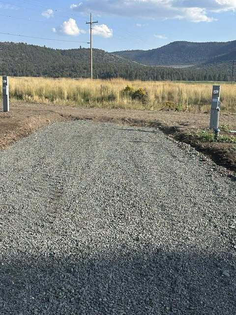 Gravel driveway leading into a field with power poles and mountains in the background under a blue sky.