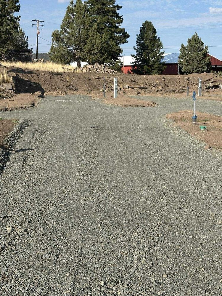 Gravel-covered construction site with small pipes, dirt, and trees under a blue sky.