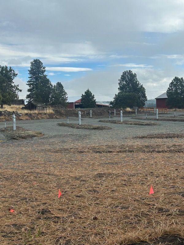 Dry field with gravel circles, white posts, red flags, and red buildings in the background under a cloudy sky.