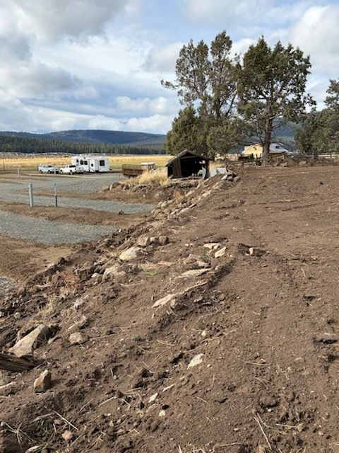Dirt embankment with rocks, leading to an open field with RVs, a small building, and trees against a cloudy sky.