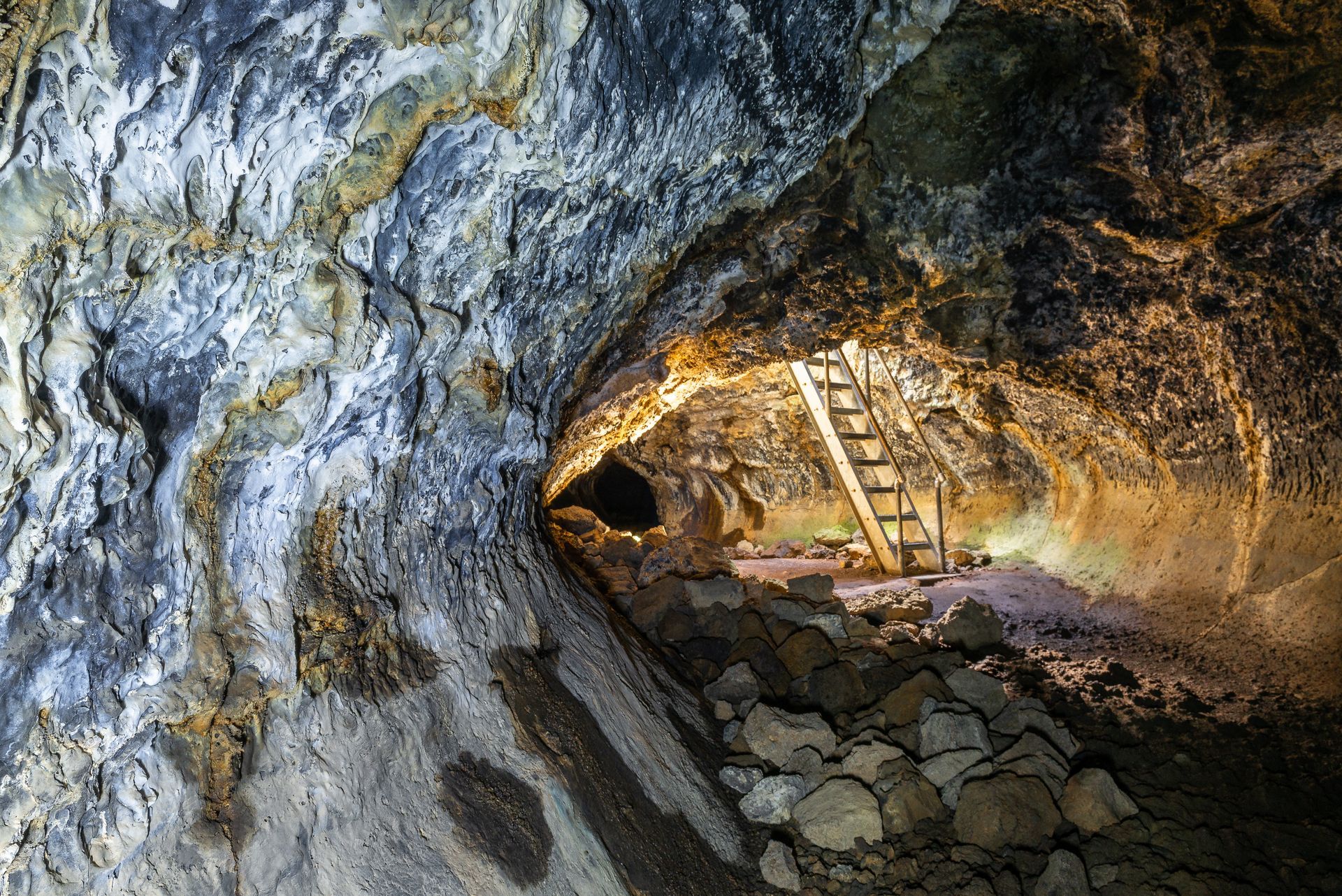 Inside a dark cave, a wooden ladder leads to the exit. Rocks and textured walls fill the space.