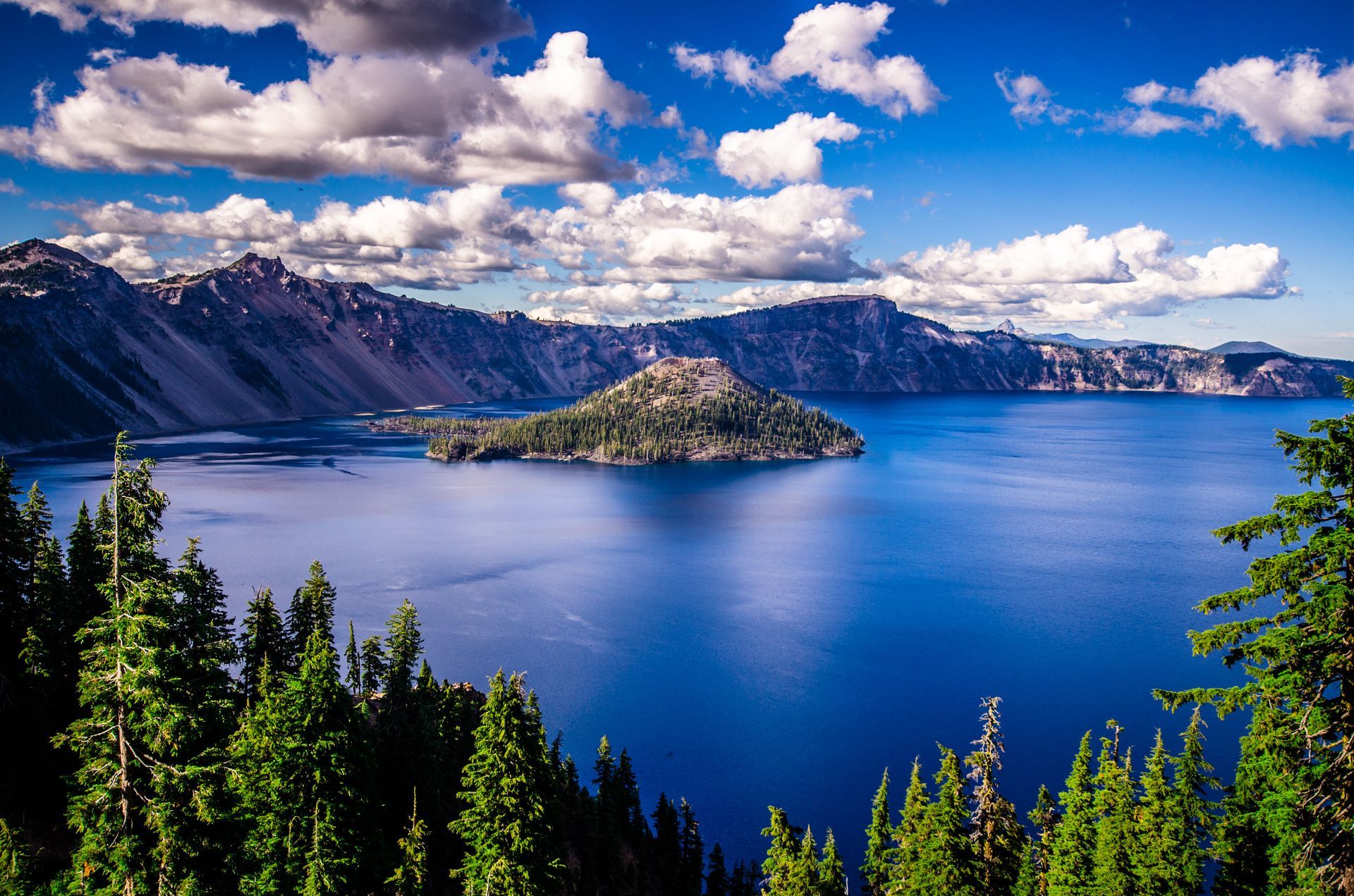 Blue lake with island, surrounded by mountains and pine trees under a bright blue sky with white clouds.