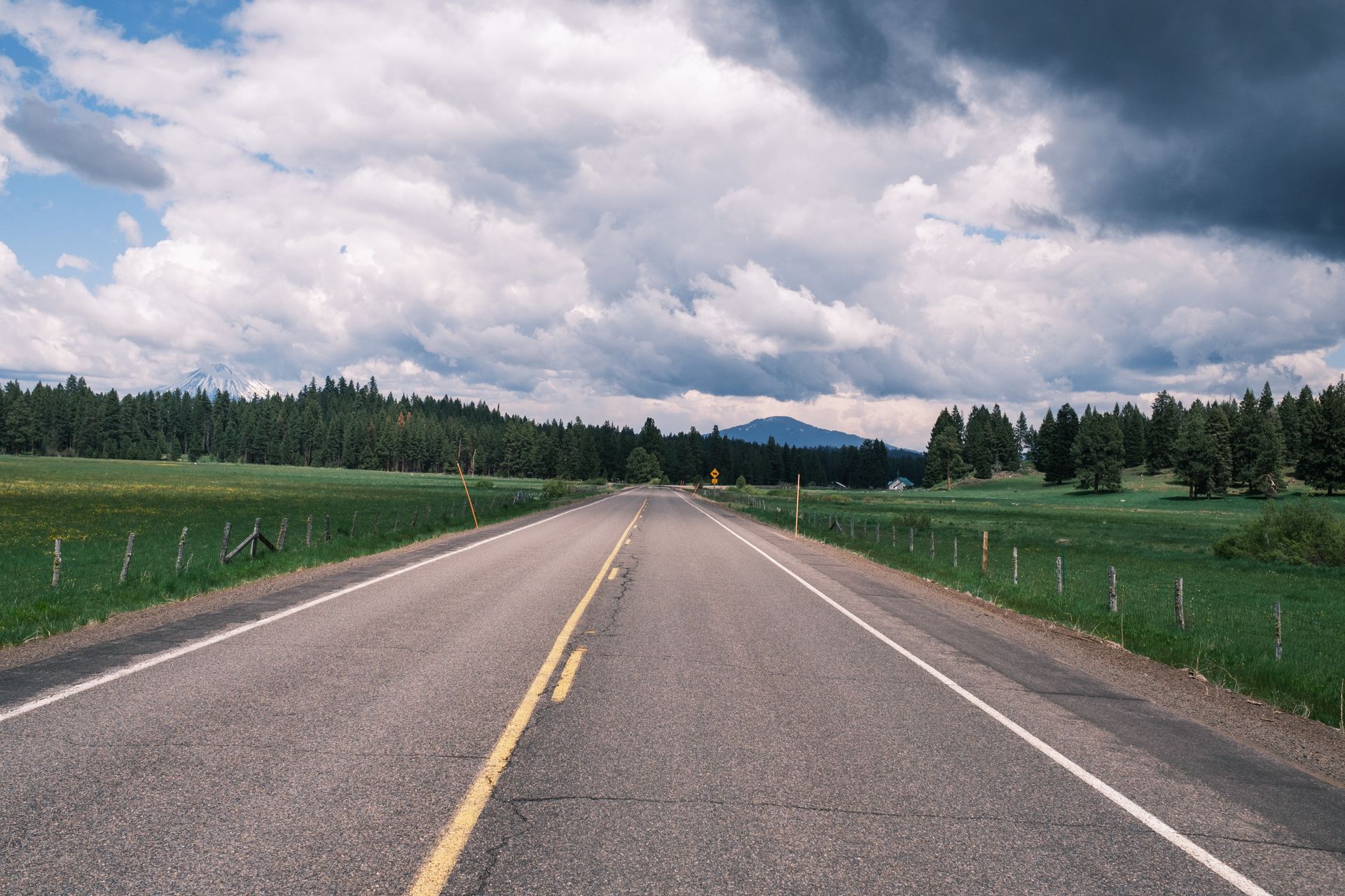 Paved road through a green field and forest under a cloudy sky.