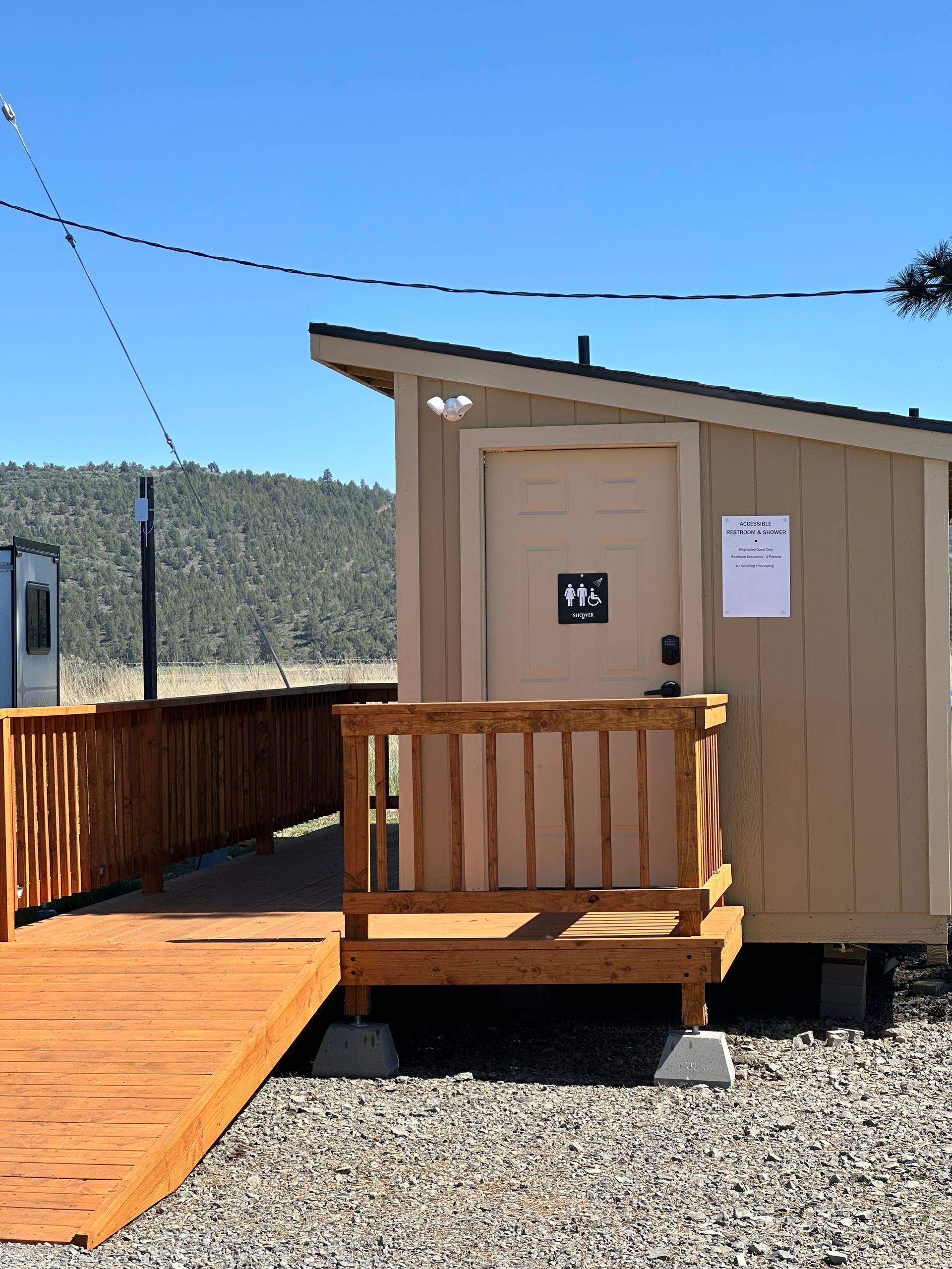 Small tan cabin with wooden porch and wheelchair ramp on a sunny gravel site.