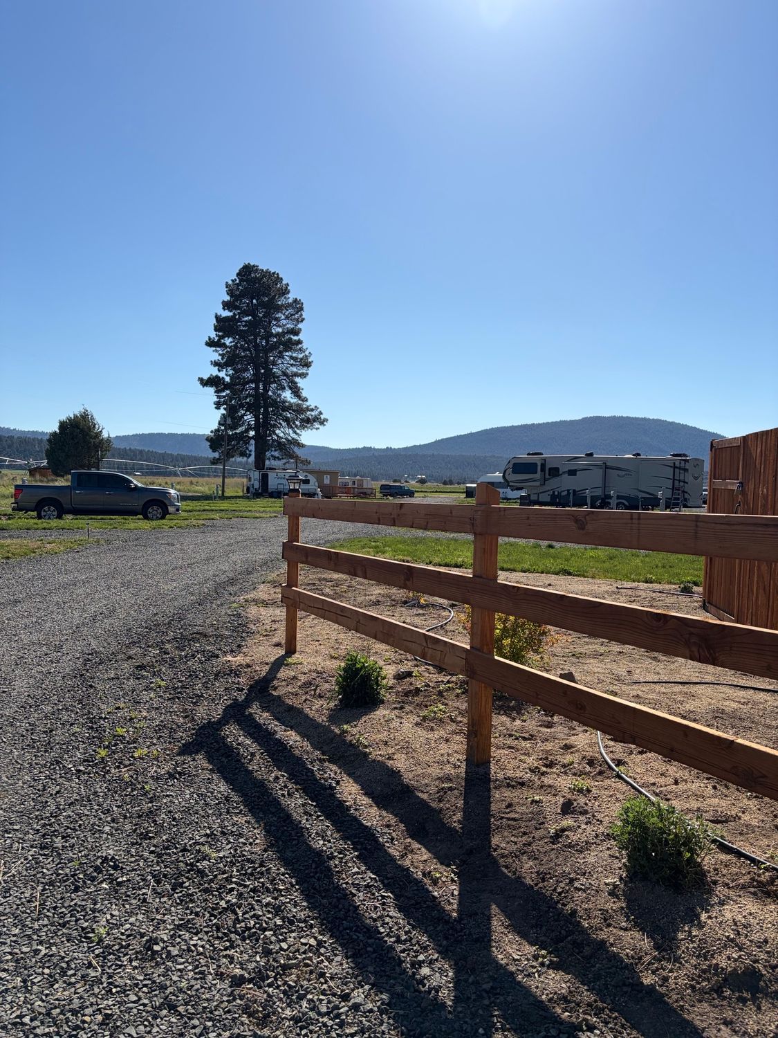 Sunny rural campsite with a wooden fence, gravel lot, RVs, and distant hills under a clear blue sky
