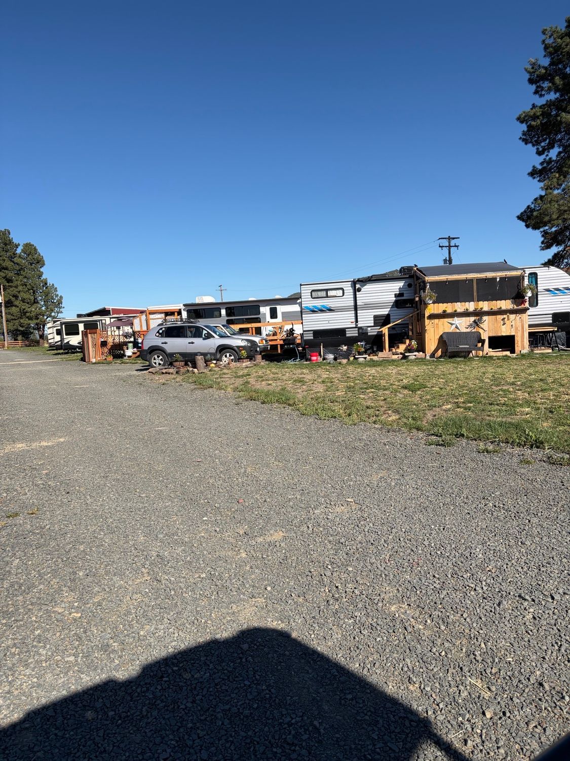 Gravel lot with parked RVs and cars under a clear blue sky, viewed from the shade of a building.