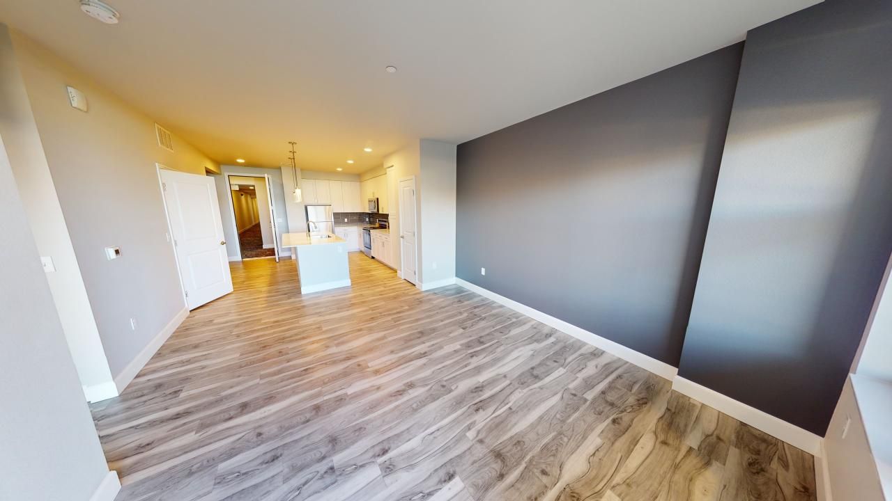 An empty living room with hardwood floors and a kitchen in the background.