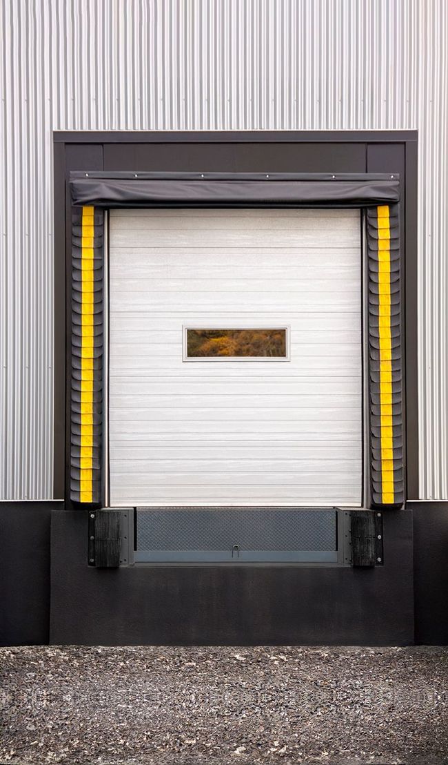 Closed white loading dock door with yellow safety bumpers in front of a metal building.