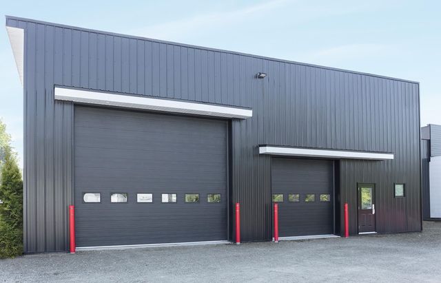Modern gray industrial warehouse with two black roll-up garage doors and red bollards