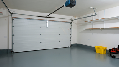 Empty garage with white roll-up door, gray floor, wall shelves, and a yellow bin on the right.
