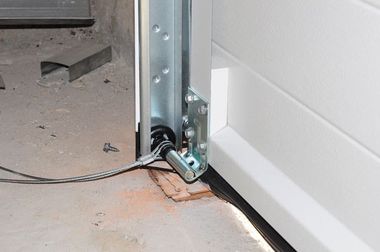 Garage door track and bottom bracket with a cable near the floor, beside a white door panel