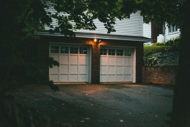 Dusk view of a double garage with lit porch lights, framed by trees and a brick wall.