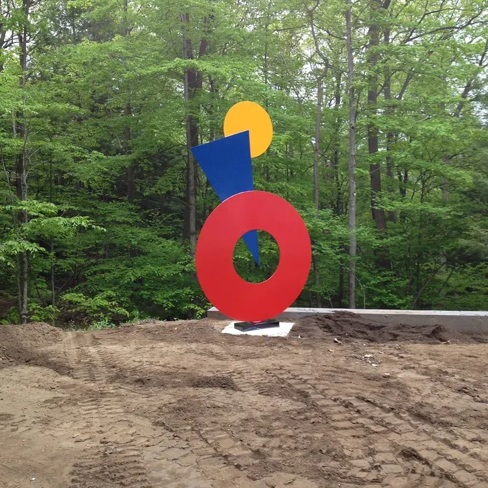 Abstract sculpture in a clearing; red circle, blue triangle, and yellow sphere; set against green trees.