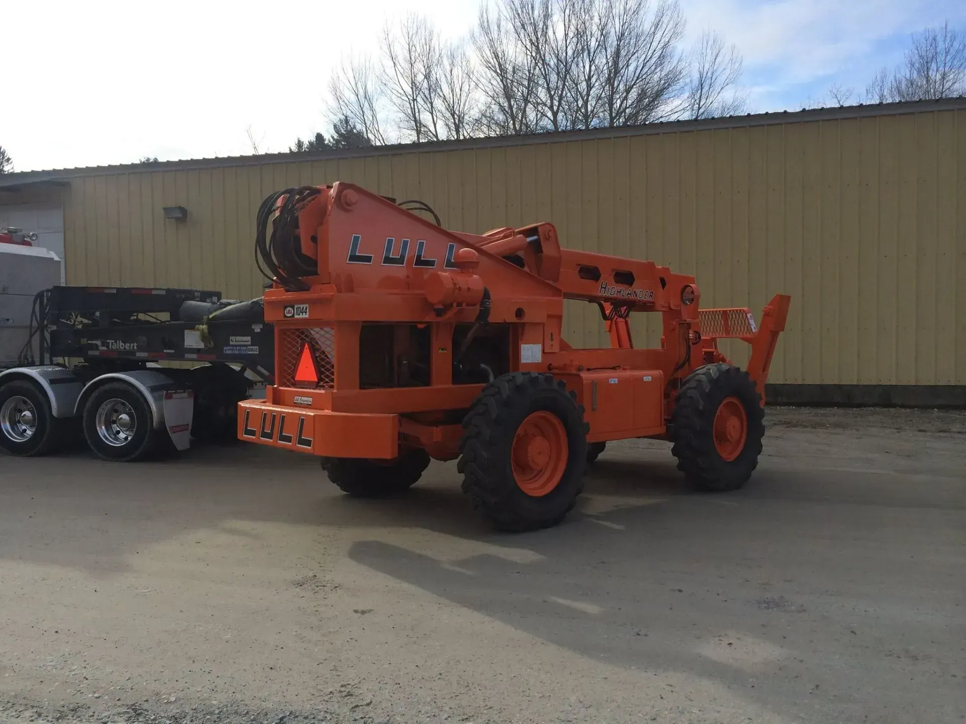 Orange Lull telehandler with black semi-truck parked in front of a tan building.