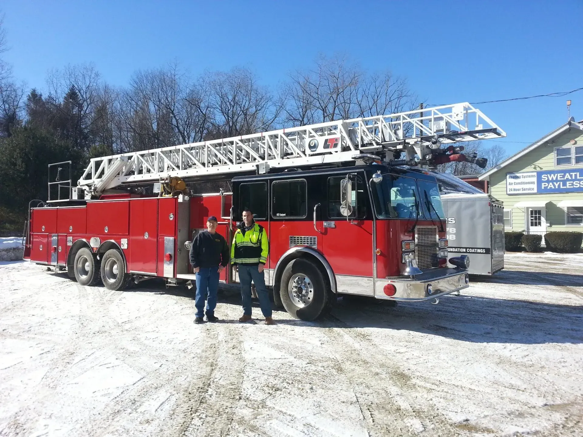 Two people stand next to a red fire truck with a ladder, parked outdoors in a snowy area.