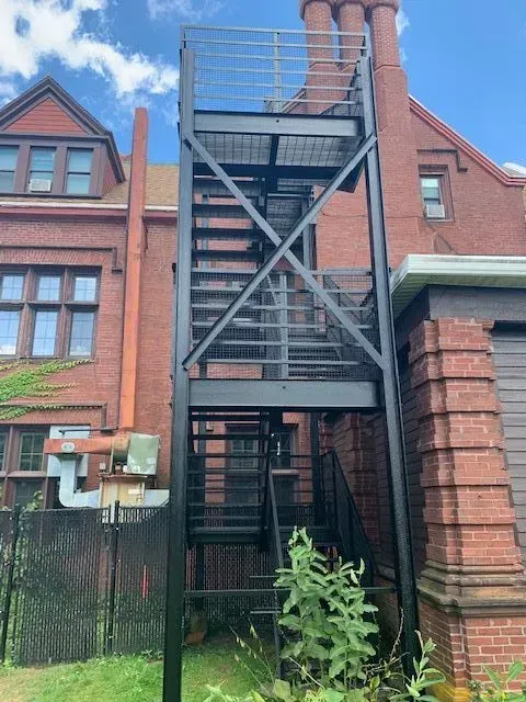 Black metal fire escape on a brick building, reaching toward the roof. Green foliage below, blue sky.