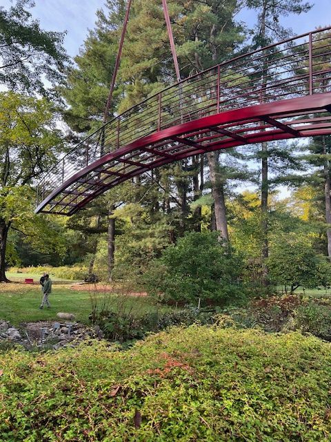 A red arched pedestrian bridge is being lifted into place over a creek in a park. Trees and foliage surround the scene.
