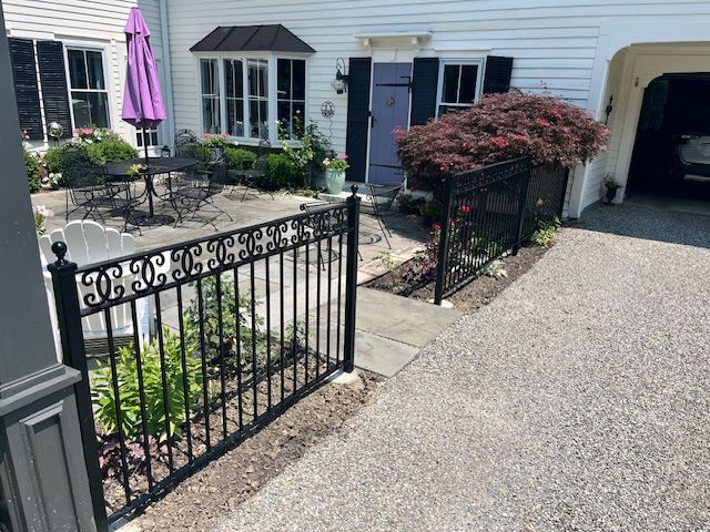 Black decorative metal fence in front of a white house with a driveway. A purple umbrella is visible.