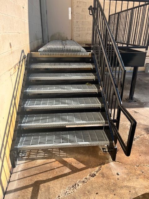 Exterior metal staircase leading to a door; black handrail; gray metal steps; concrete ground.