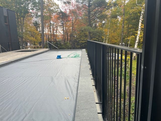Black railing along a deck overlooking a covered pool in a wooded area with fall foliage.