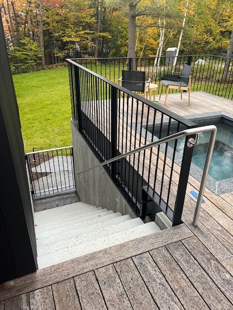 Staircase leading down to a hot tub. Black railing with stainless steel handrail, wooden deck, green lawn, trees.