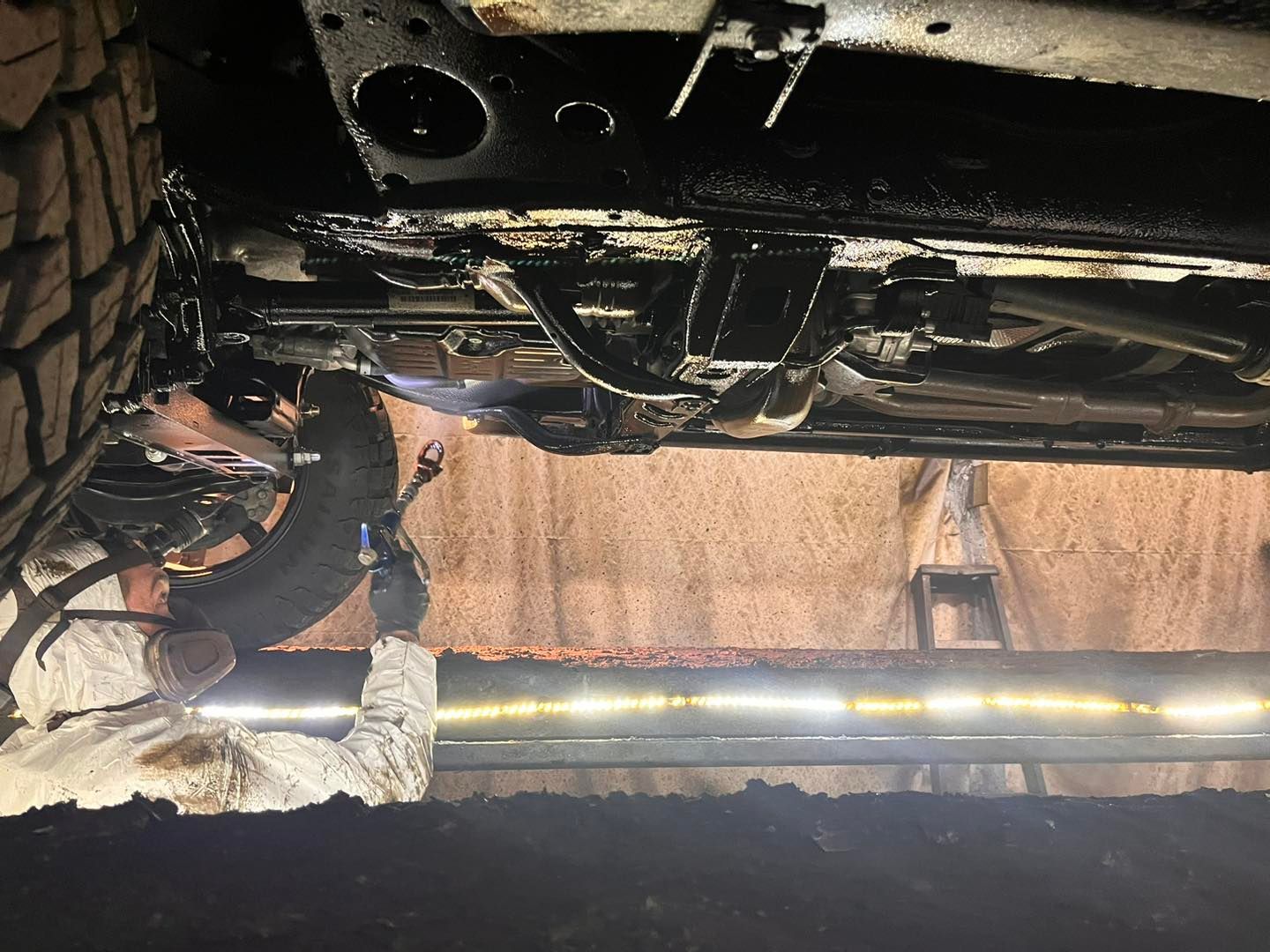 Person spraying black coating under a vehicle. Workshop setting, low angle shot.