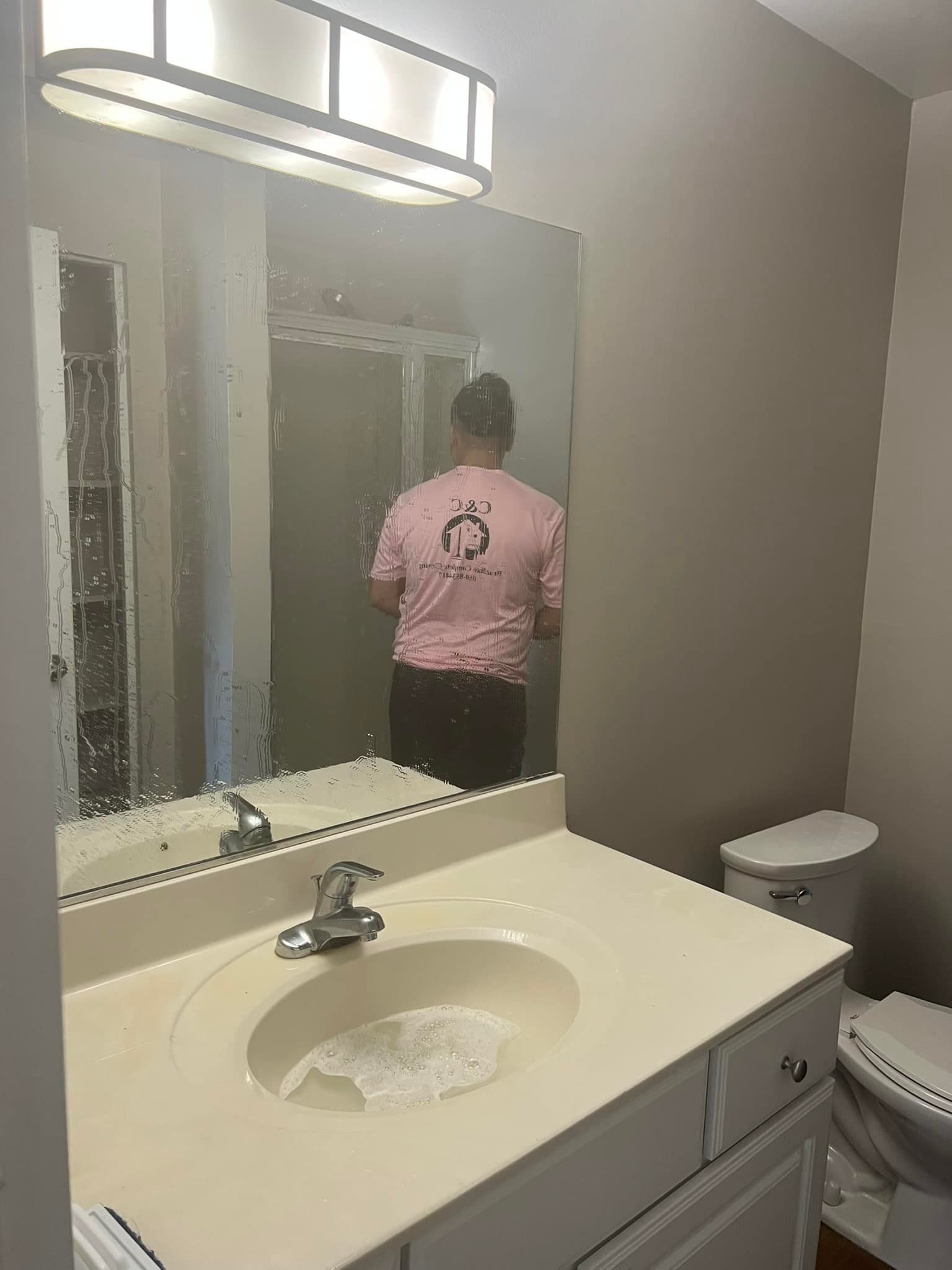 A man in a pink shirt is standing in front of a bathroom sink and mirror.