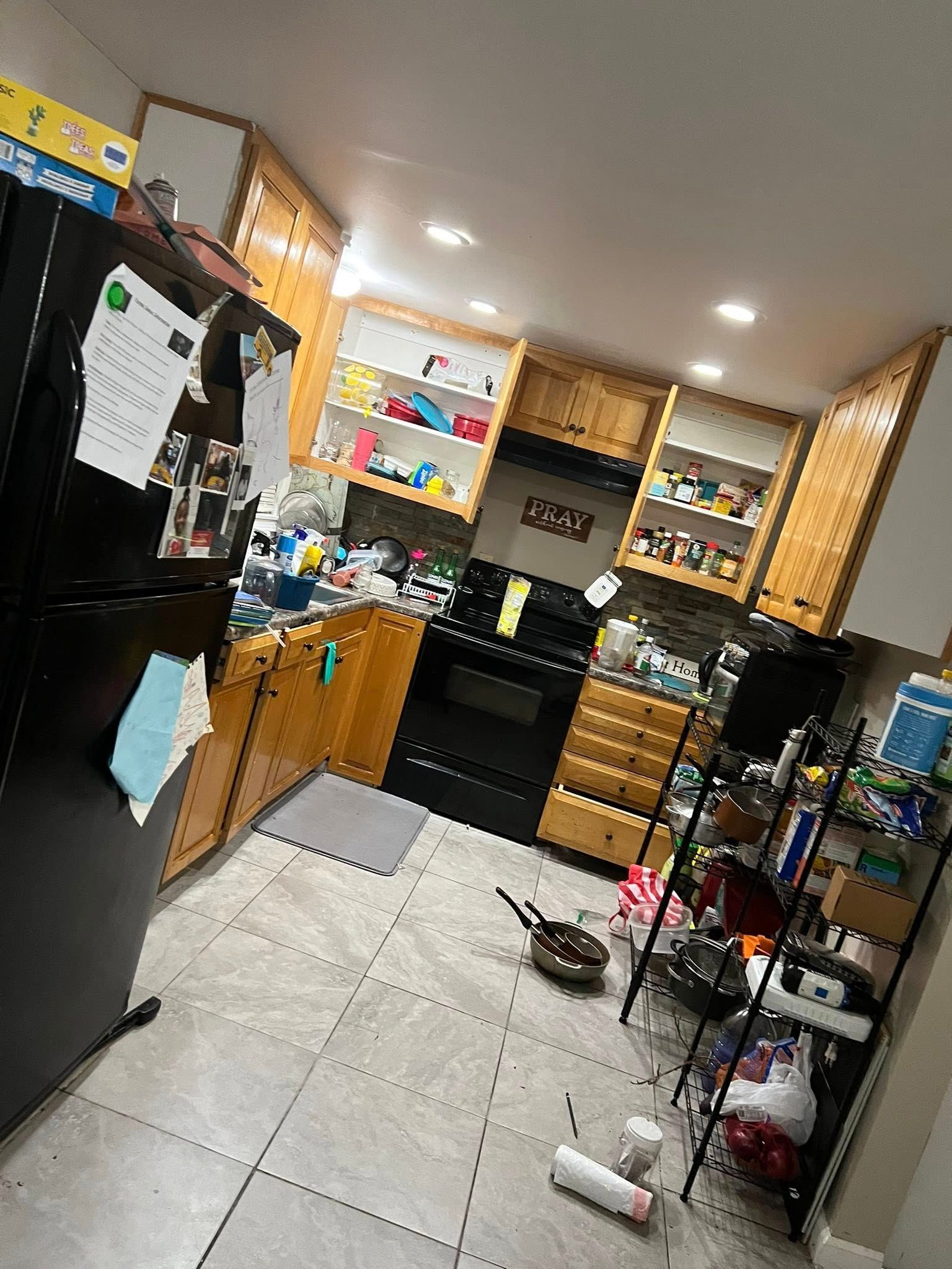A kitchen with a black refrigerator , wooden cabinets , a stove , and a pan on the floor.