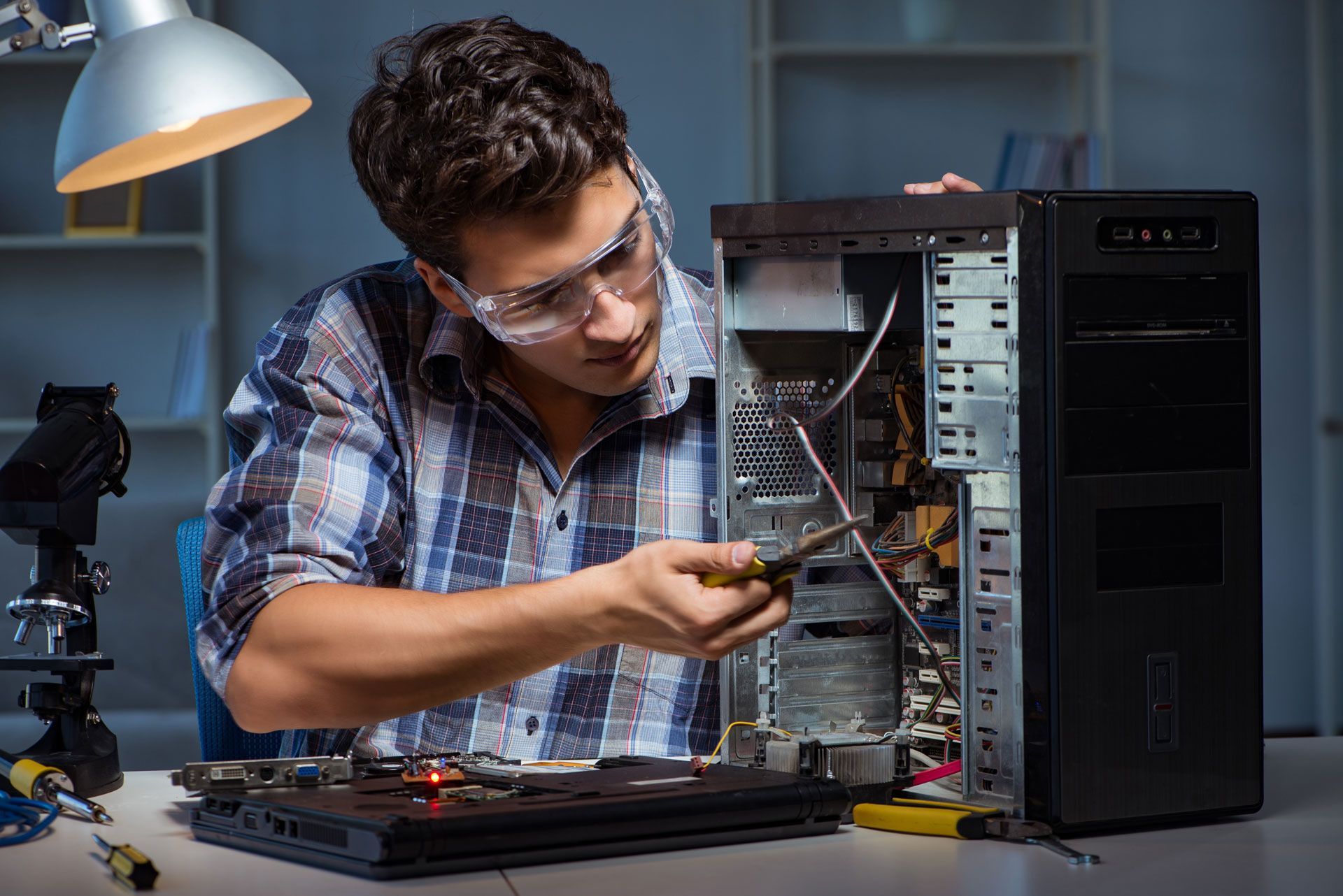 Man in safety glasses repairs computer tower with a screwdriver in a well-lit workshop.