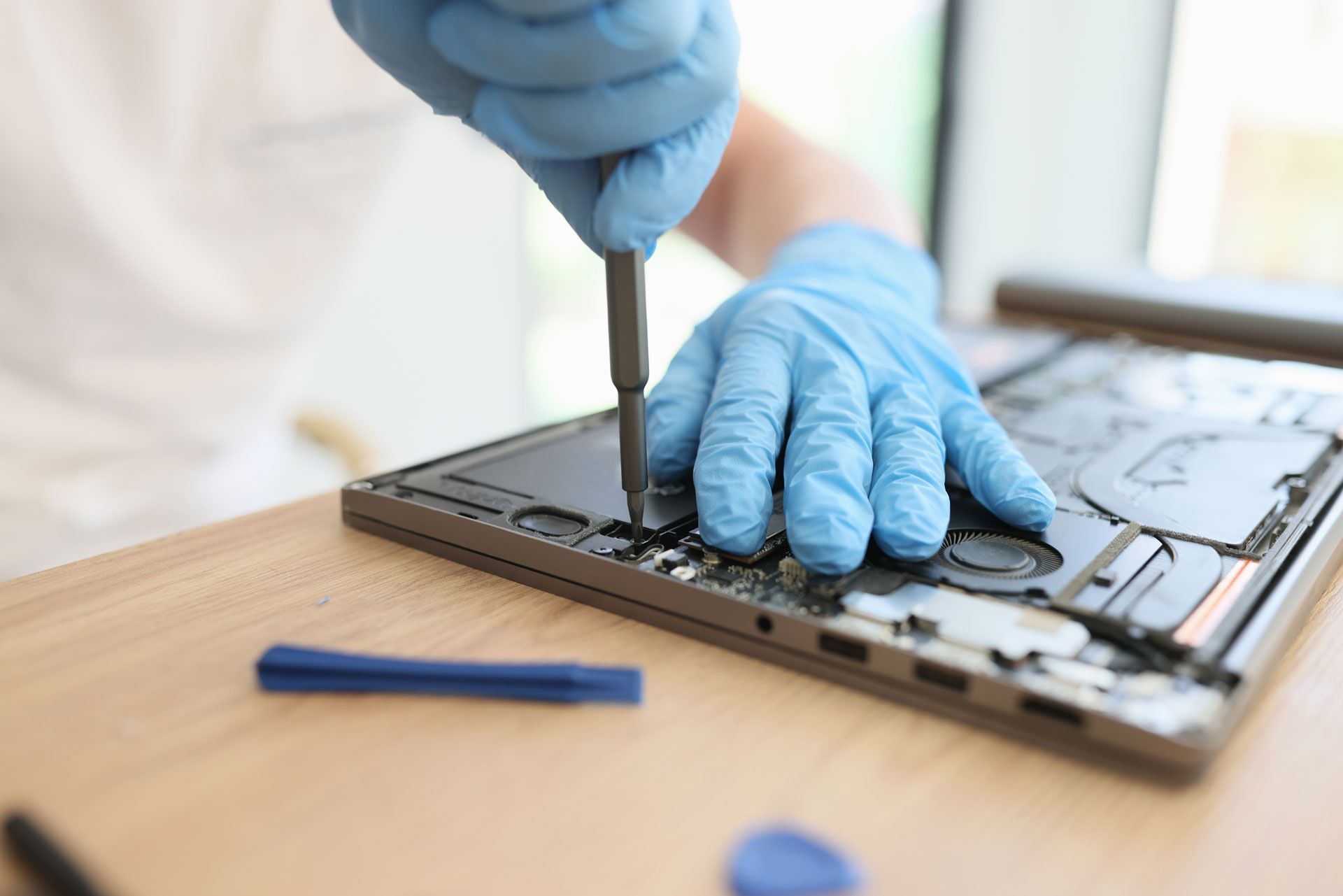 Person in blue gloves using a screwdriver to repair a laptop on a wooden surface.