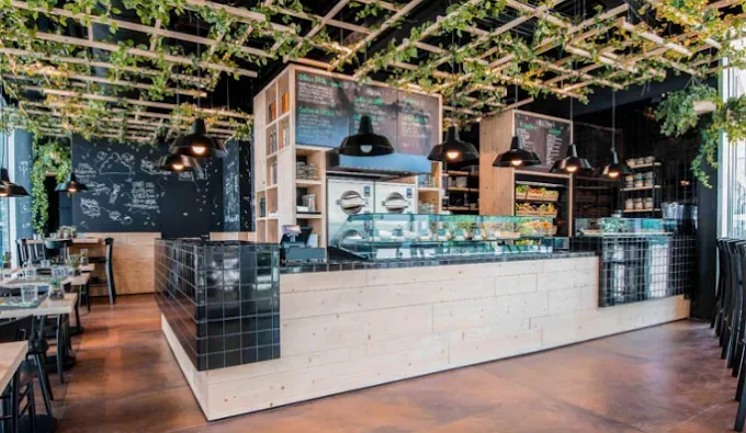 Interno del bar con legno chiaro e dettagli neri, bancone con esposizione di cibo, soffitto decorato di verde, tavoli e sedie.