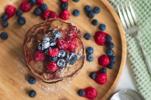 Pila di pancake guarniti con lamponi e mirtilli, serviti su un piatto di legno con forchetta e cucchiaio.