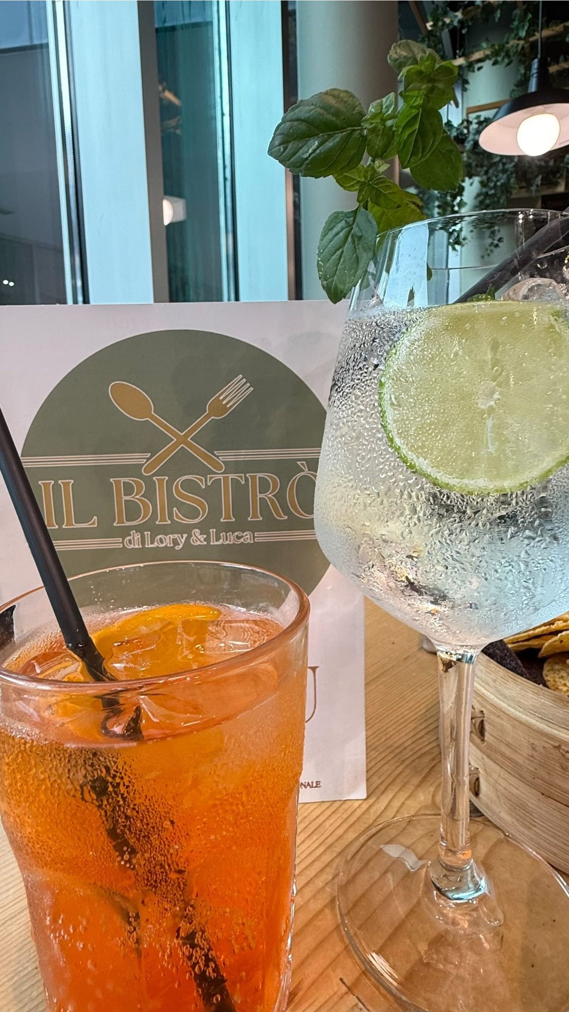 Due cocktail su un tavolo: uno all'arancia con cannuccia, uno trasparente con lime e menta. Sullo sfondo, l'insegna di un bistrot.