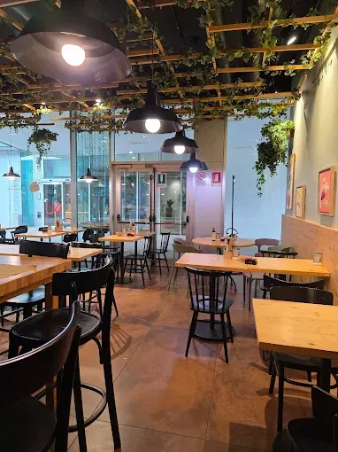 Interno di un ristorante con tavoli in legno e sedie nere. Luci zenitali e fogliame verde decorano l'ambiente.