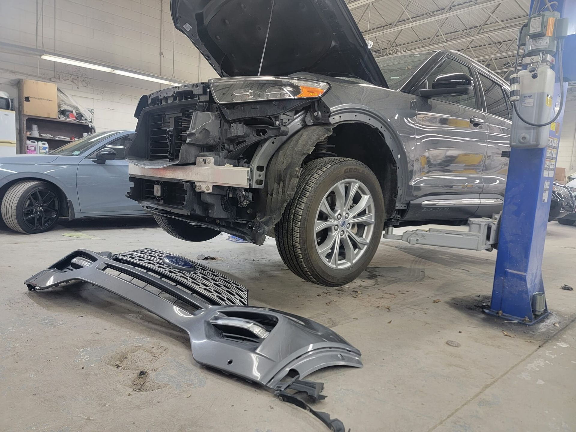 Car in repair shop, front end damaged with detached bumper. Grey SUV elevated on lift.
