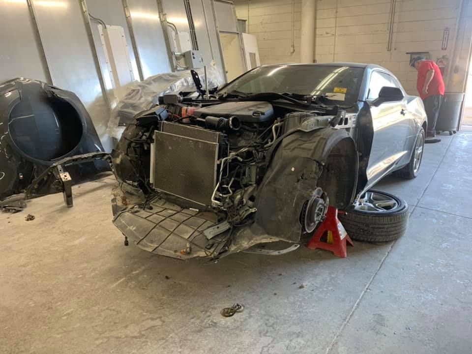 Damaged gray car on jack stands in a repair shop, front end severely crumpled.