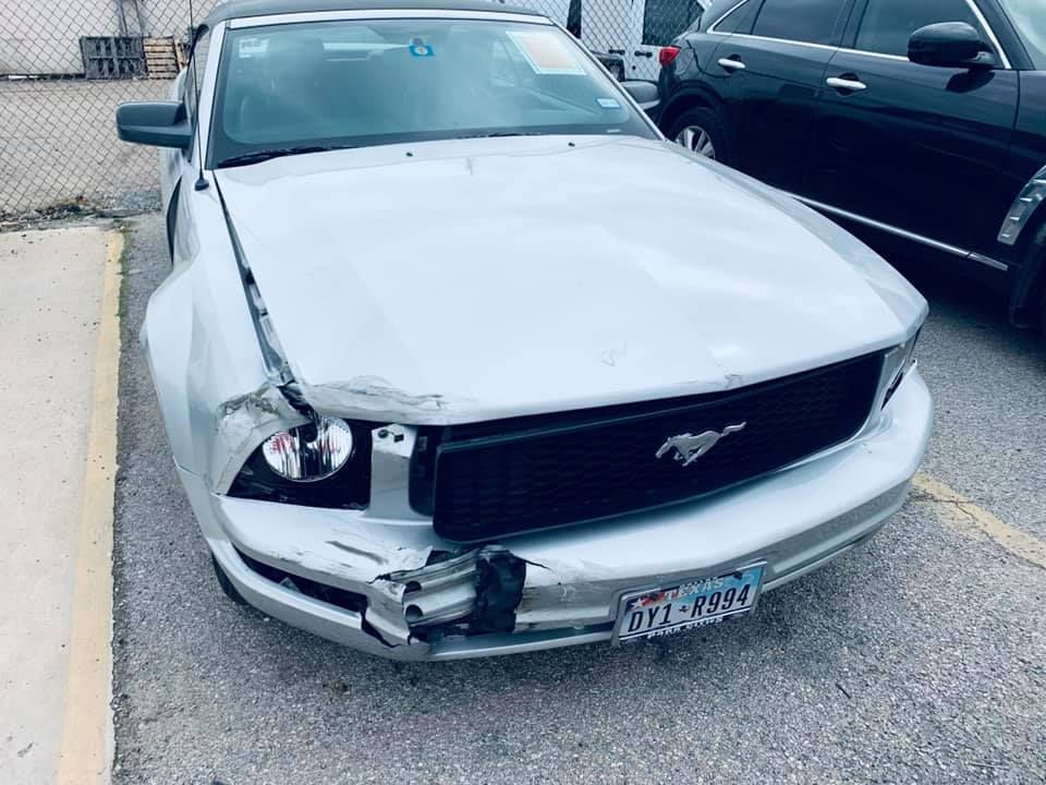 Silver Ford Mustang convertible with front-end damage, parked outdoors, black grille, Texas license plate.