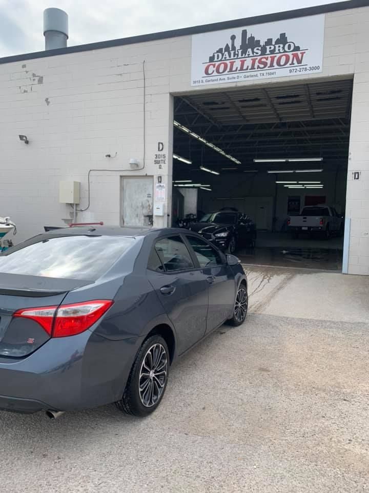 Gray car parked outside a collision repair shop with open garage door; other vehicles visible inside.