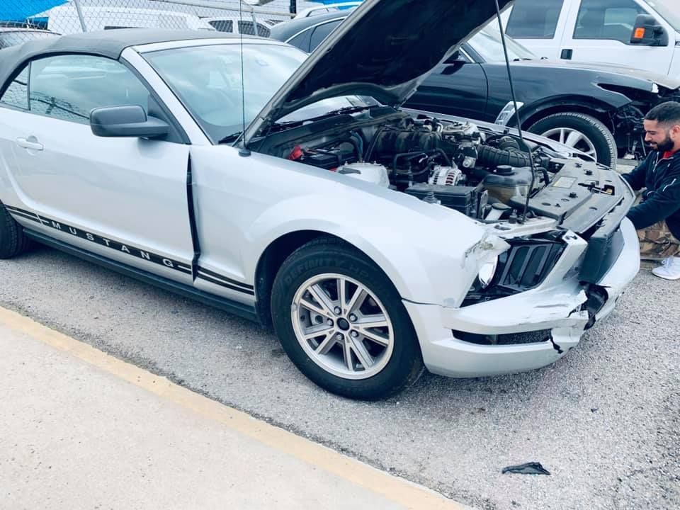 Silver Mustang convertible with front-end damage, hood open, man inspecting it outdoors.