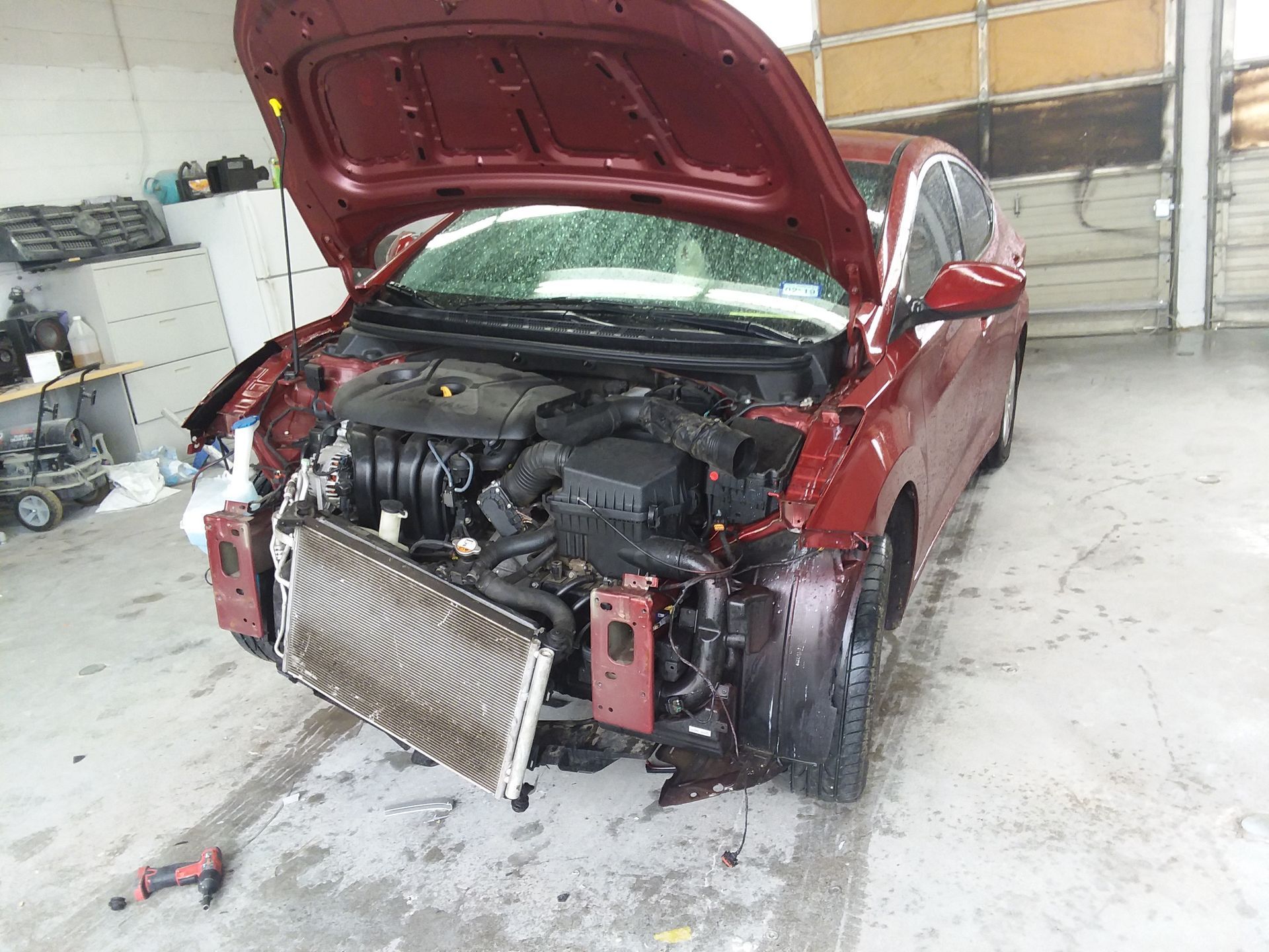 Red car with open hood, front-end damage in repair shop.